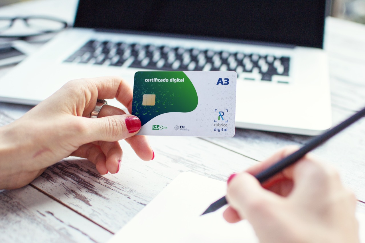 Cartão certificado digital A3 da Rubrica Digital sendo segurado sobre teclado de laptop, com selos ICP-Brasil e ITI visíveis