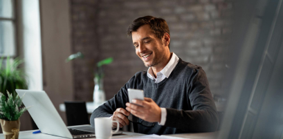 Pessoa sorrindo enquanto usa celular e laptop em mesa de escritório - uso típico do certificado digital e-CPF (imagem oficial Soluti)